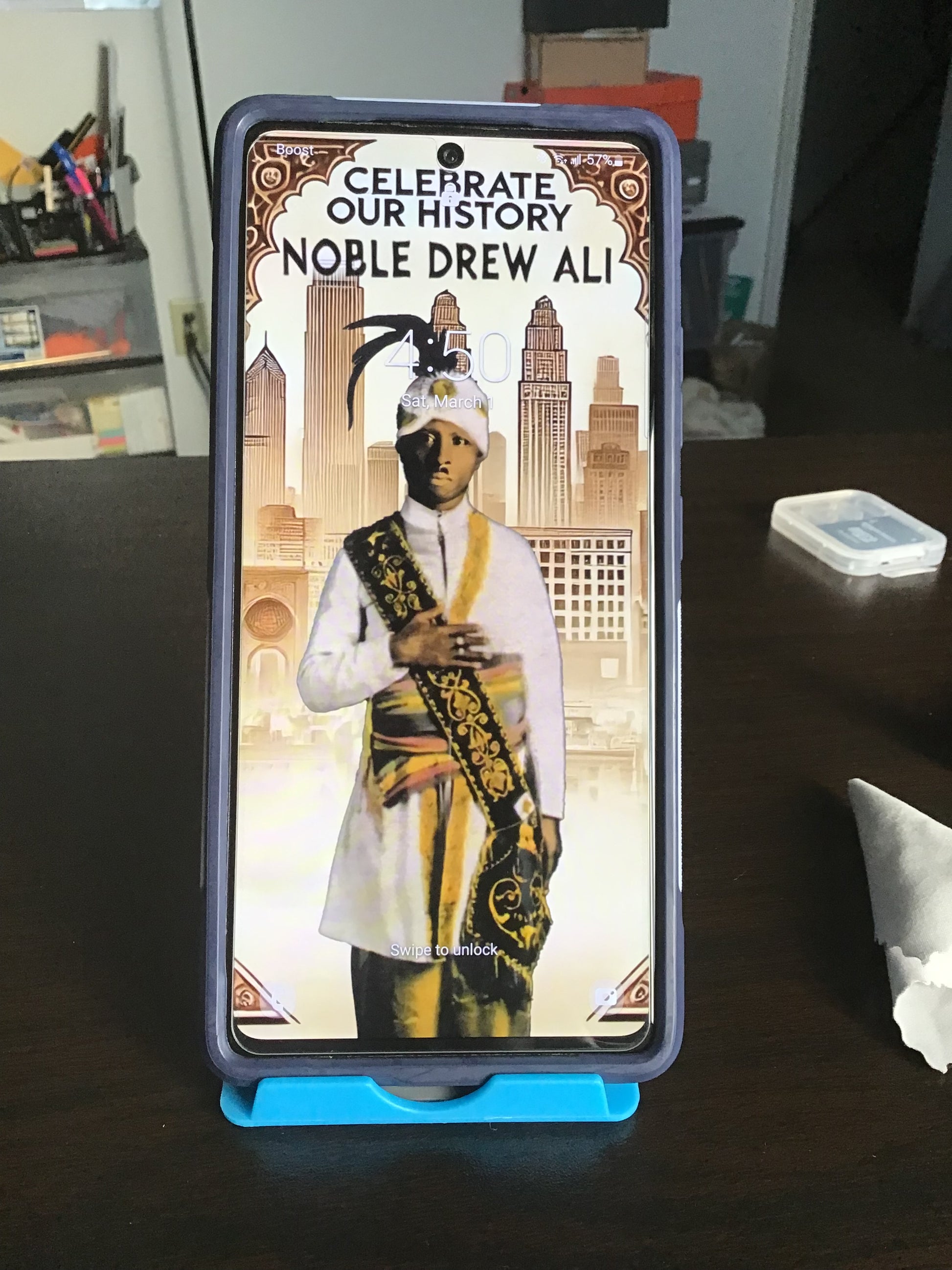 A smartphone in a dark protective case stands on a blue stand on a dark wooden table. The screen displays a custom graphic with the text 'CELEBRATE OUR HISTORY NOBLE DREW ALI' and an illustration of a historical figure in a white and gold robe.